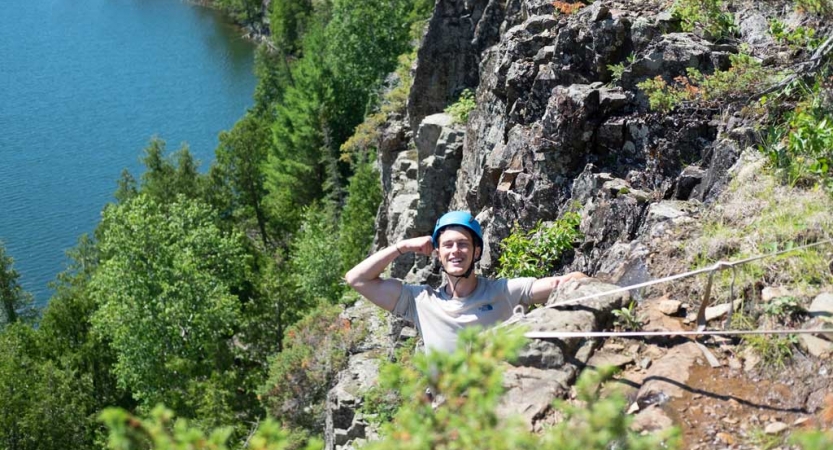 A person wearing safety gear and secured by ropes stands just below the edge of a rocky cliff, smiles and salutes the camera. They are above a blue body of water. 